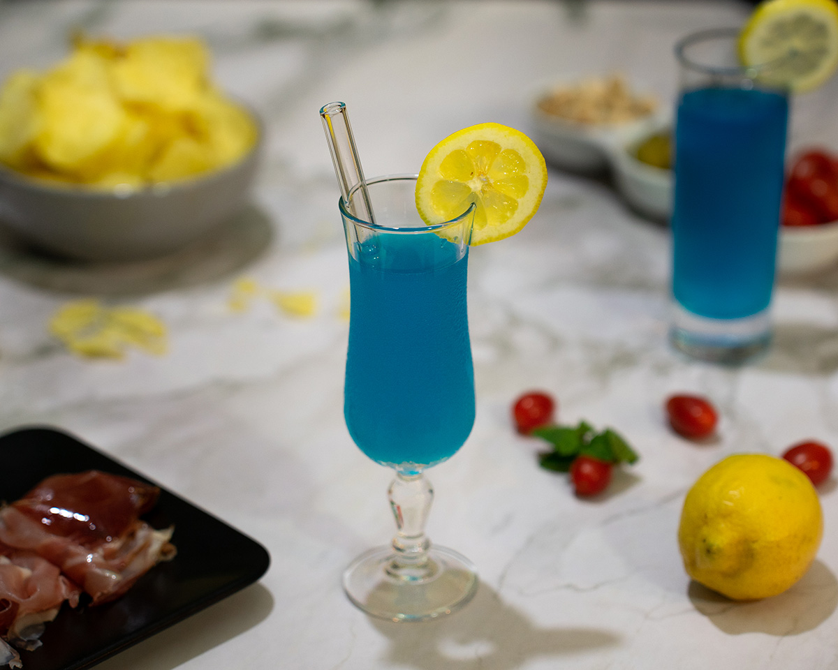 Photo culinaire d’un cocktail blue lagoon accompagné de jambon de pays et de chips sur une table d’apéritif, idéale pour illustration de bar ou restaurant.