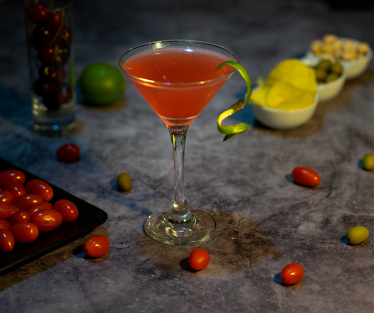 Photo culinaire d’un cocktail Cosmopolitan accompagné de tomates cerises et de chips sur une table d’apéritif, idéale pour illustration de bar ou restaurant.