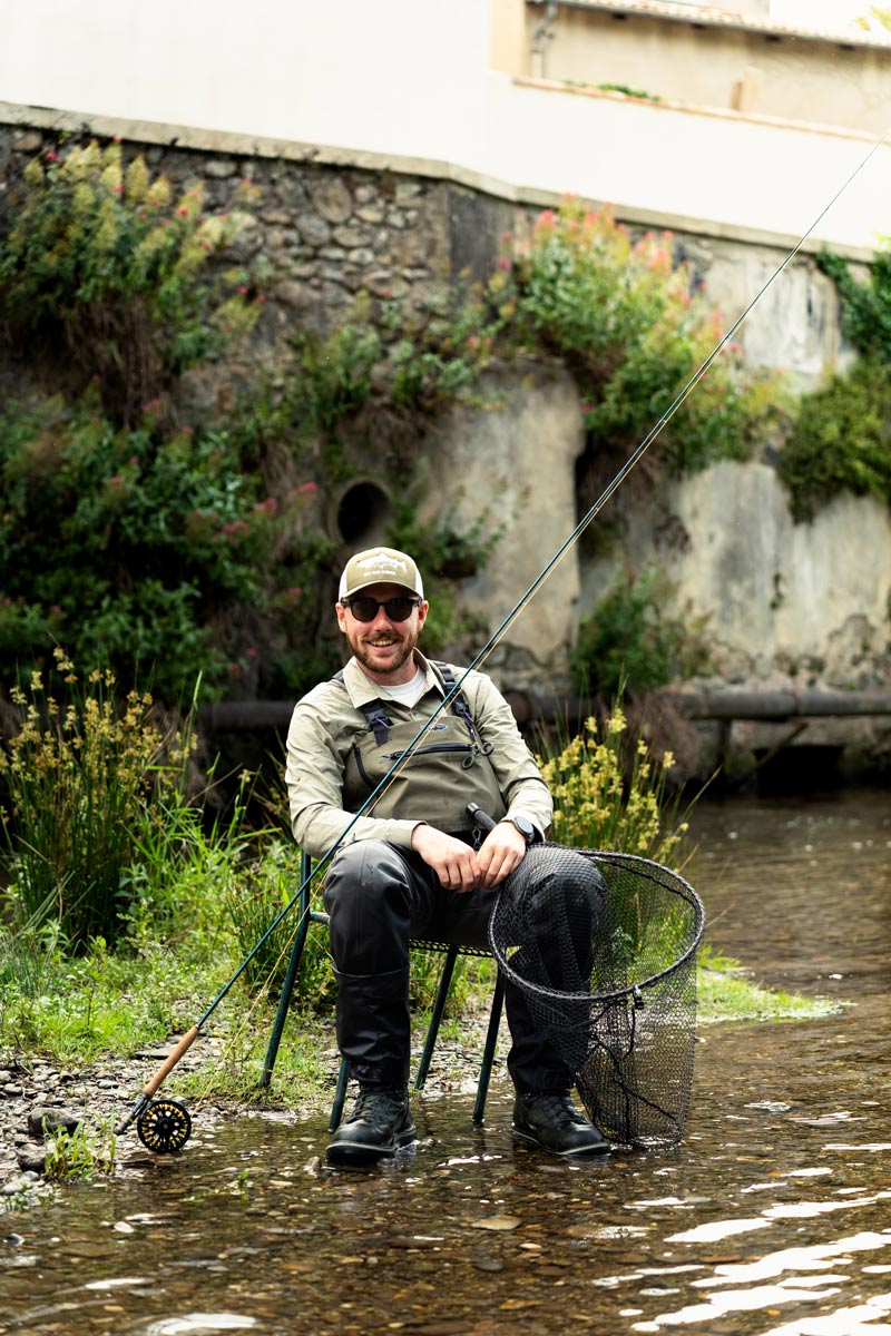 Photo de Maxime Tourrisseau équipé d'une canne à mouche et d'une épuisette, assis sur une chaise au milieu de la rivière